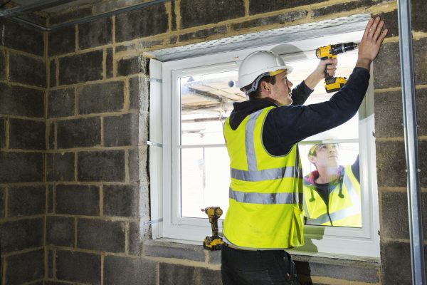 A workman on a construction site, a builder in hard hat using an electric drill on a window sill.